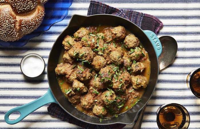 Meatballs with sliced parsley in cast iron skillet, seeded challah atop striped tablecloth.