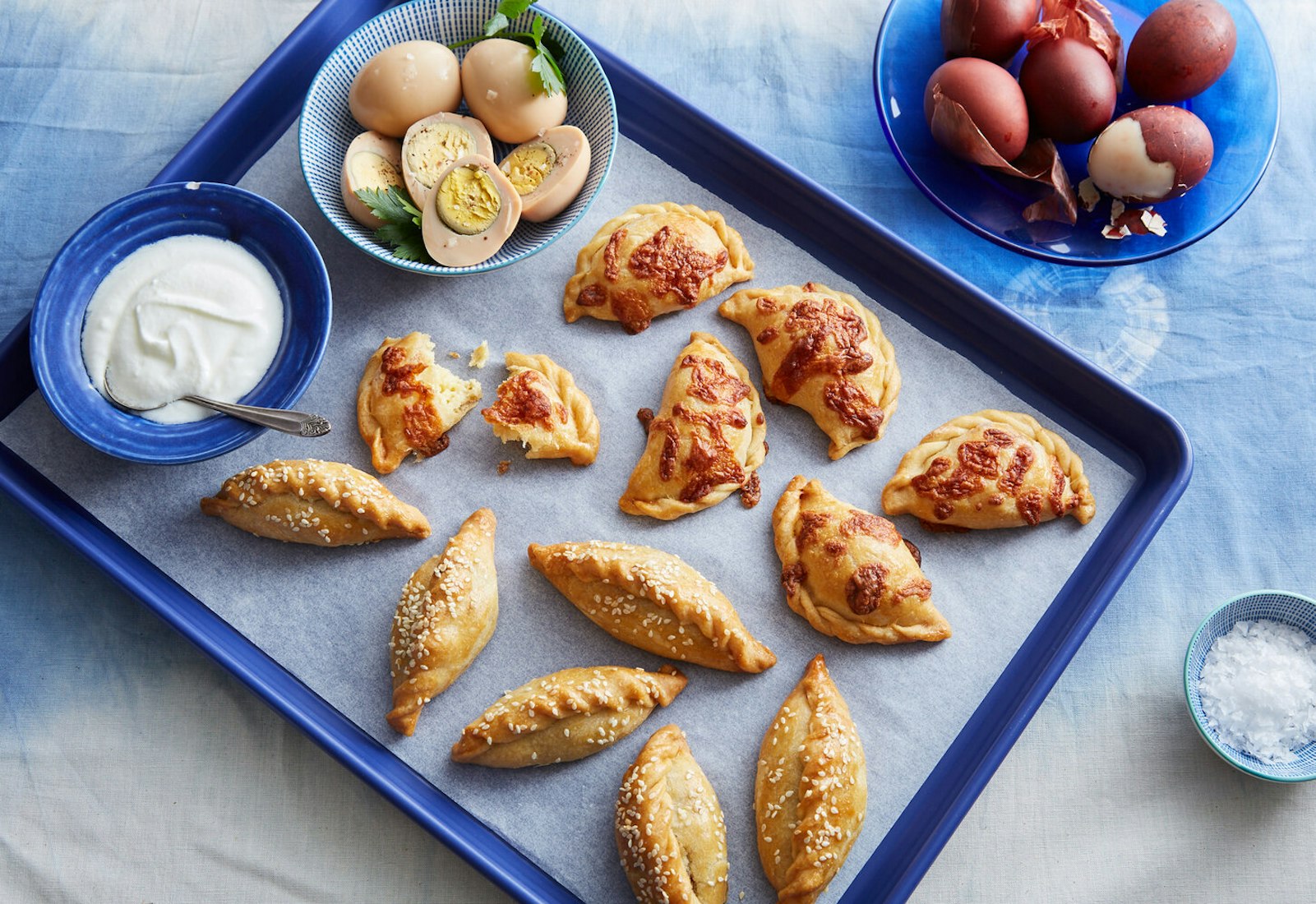 Borekitas topped with cheese and sesame seeds with side dishes of hard boiled eggs and yogurt atop blue tablecloth.