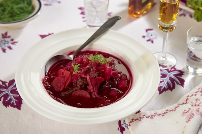 Borscht with sprigs of dill alongside liquor in champagne flute atop purple and white tablecloth.