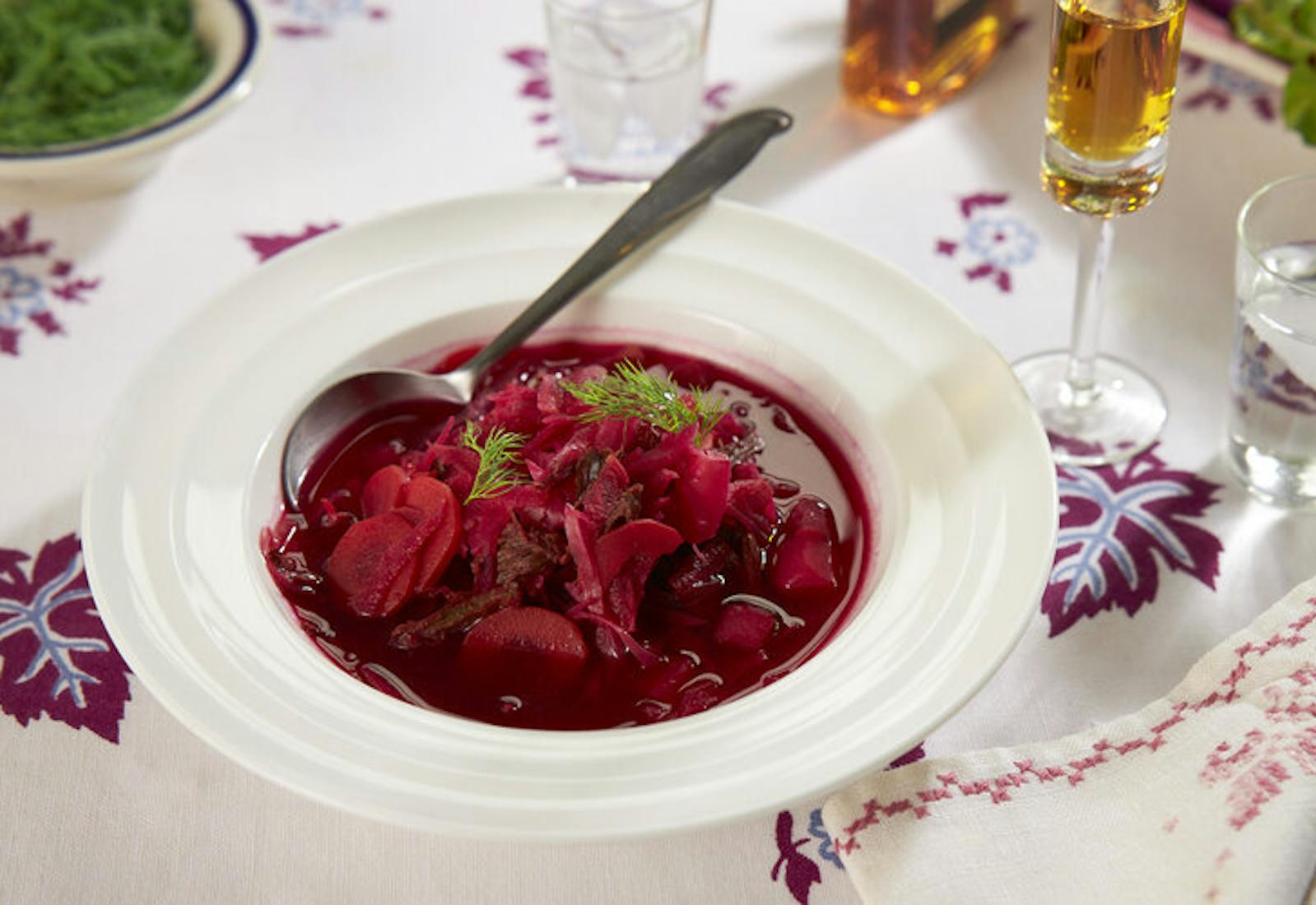 Borscht with sprigs of dill alongside liquor in champagne flute atop purple and white tablecloth.