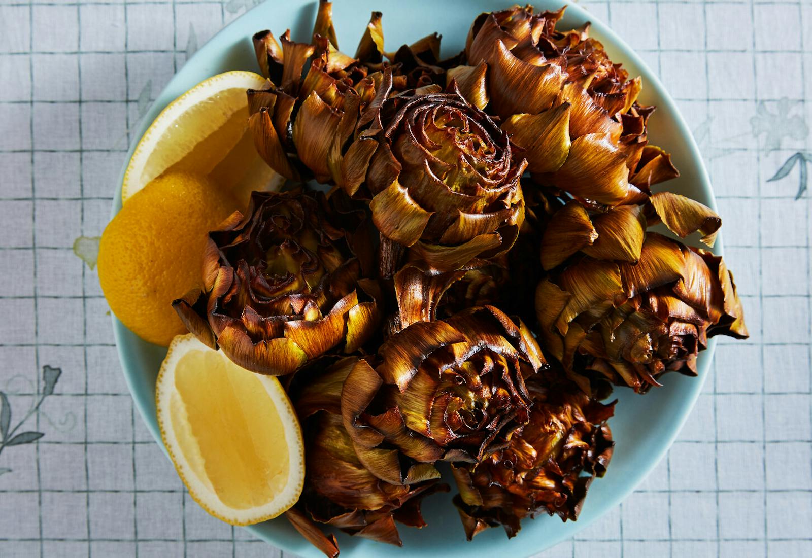 Fried artichokes with lemon wedges in blue bowl, atop grid-patterned tablecloth.