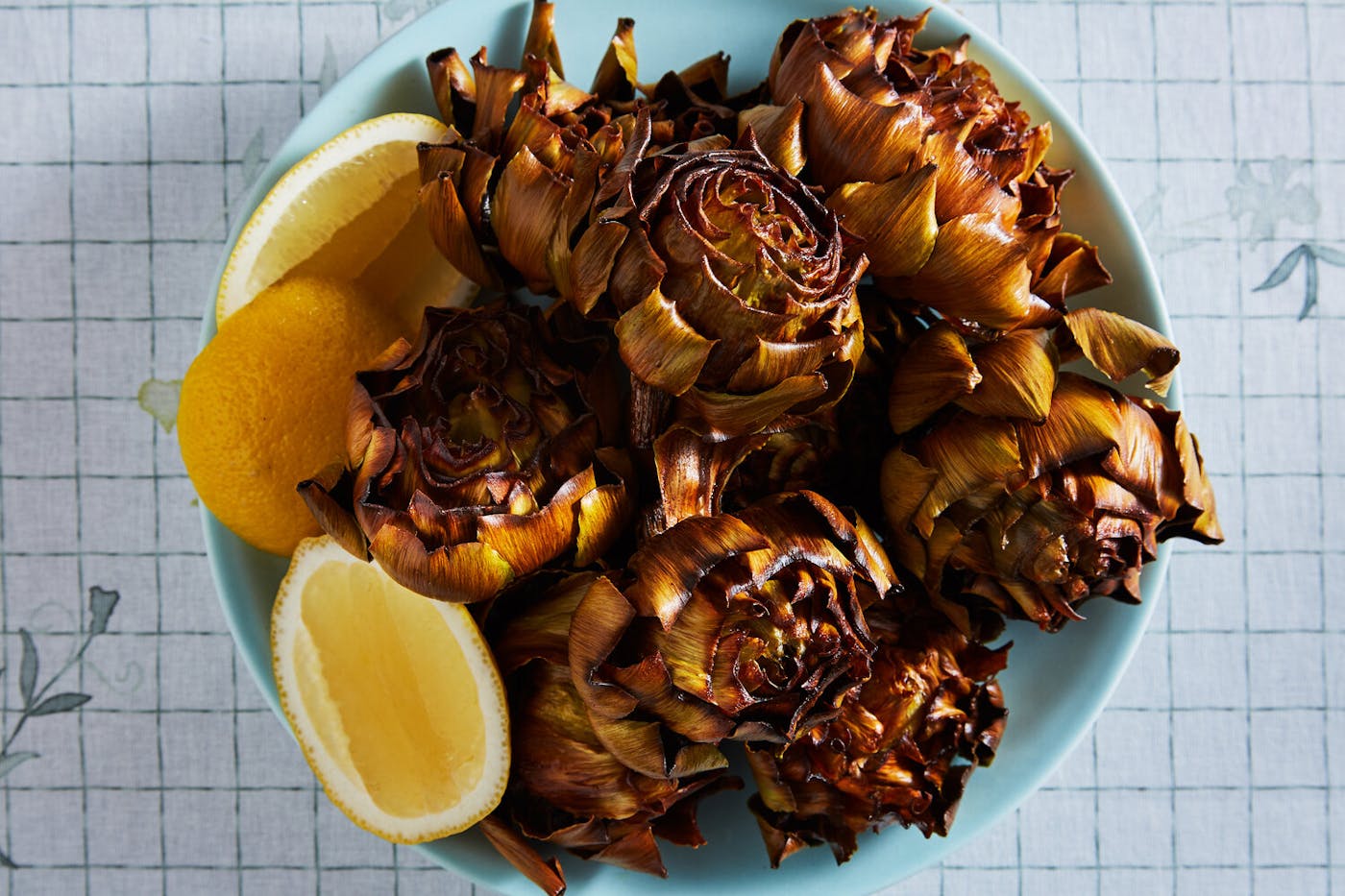 Fried artichokes with lemon wedges in blue bowl, atop grid-patterned tablecloth.