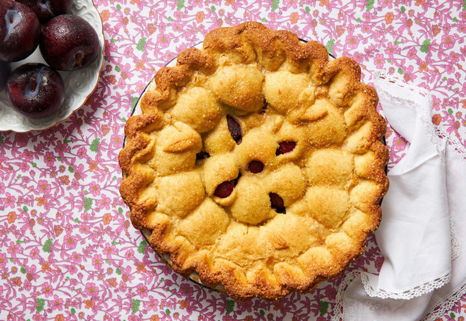Plum cake with golden crust, plate of plums atop pink floral tablecloth.