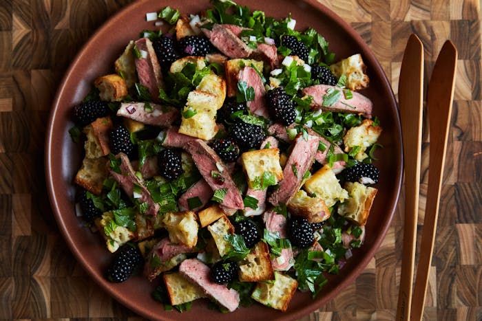 Steak and blackberry salad garnished with chopped mint on red plate with wooden tongs atop wooden surface.