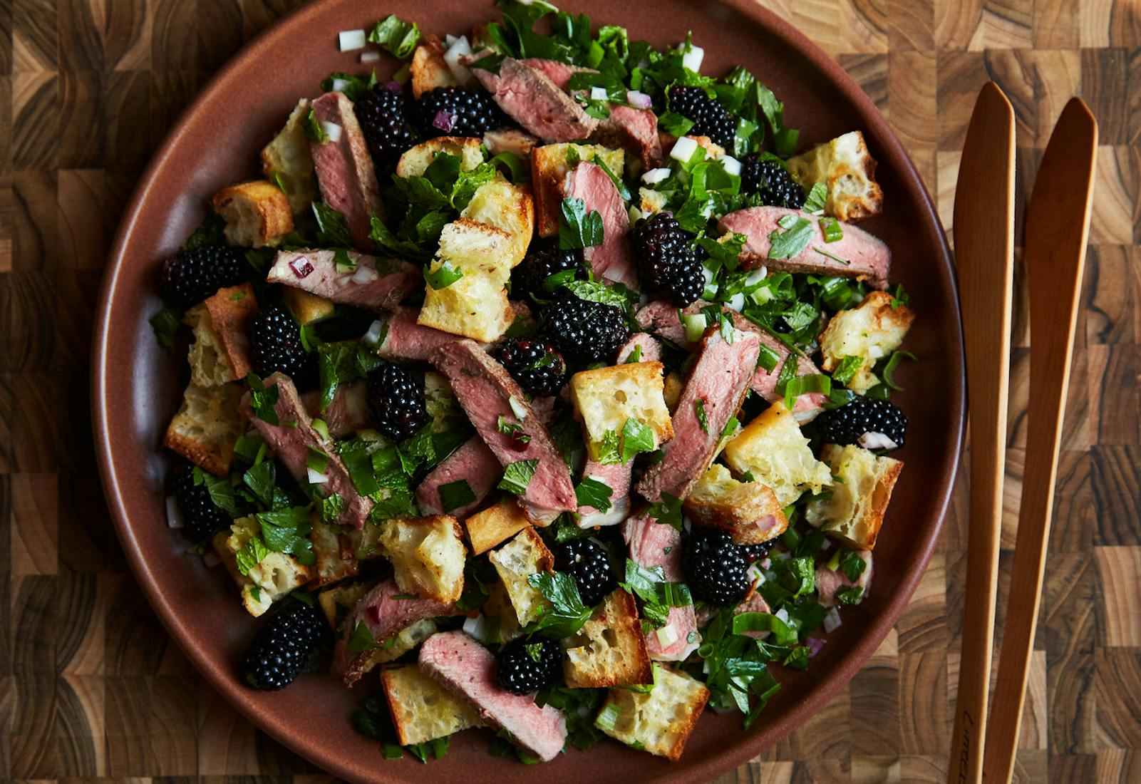 Steak and blackberry salad garnished with chopped mint on red plate with wooden tongs atop wooden surface.