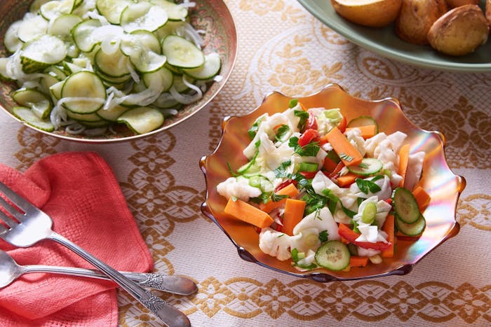 Fermented vegetable salad in orange scalloped dish alongside bowls of roasted potatoes and cucumber salad, atop orange patterned tablecloth.