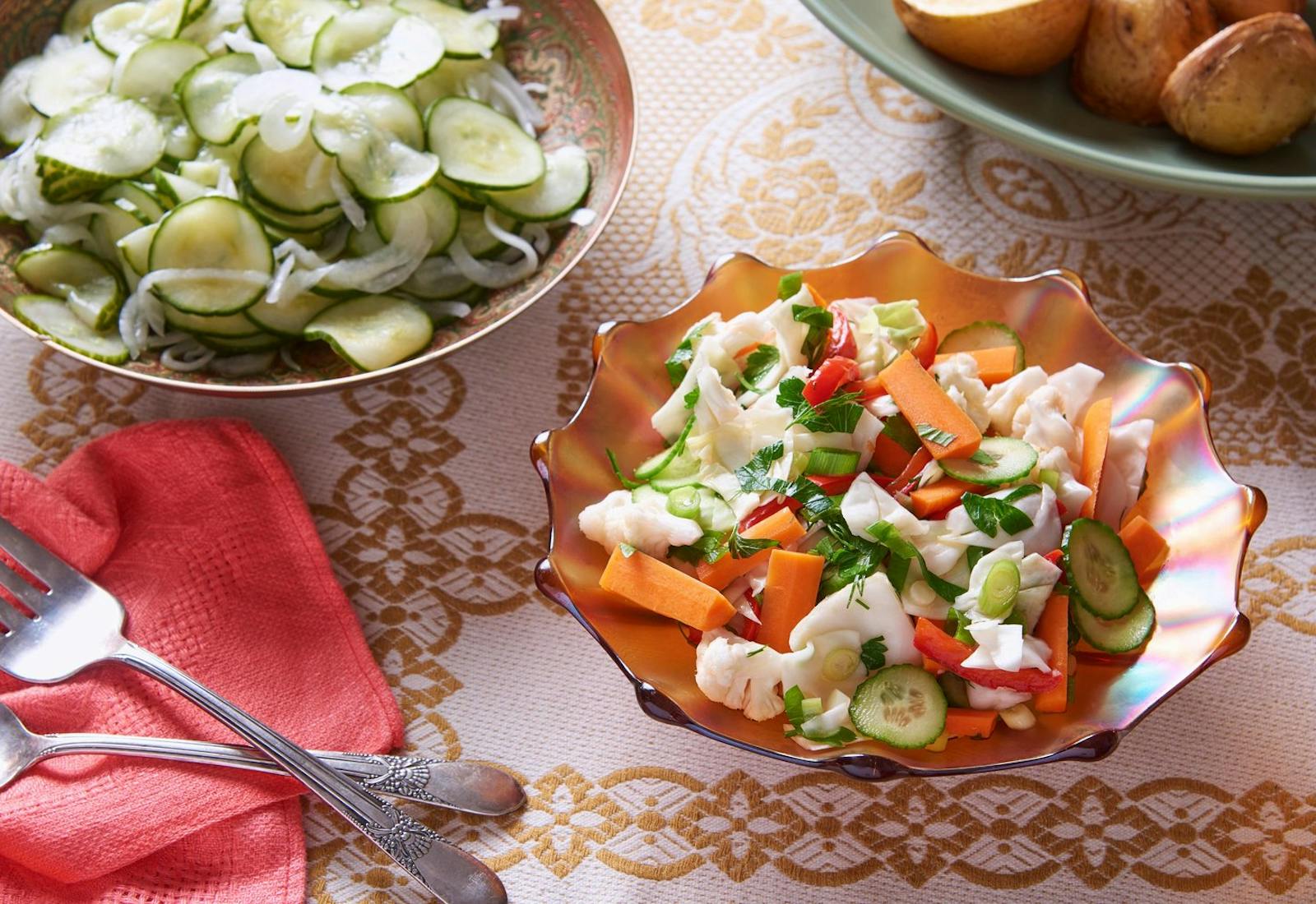 Fermented vegetable salad in orange scalloped dish alongside bowls of roasted potatoes and cucumber salad, atop orange patterned tablecloth.