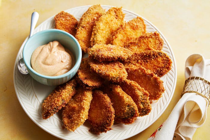 Fried zucchini chips with dish of smoked paprika aioli on white plate atop yellow surface.