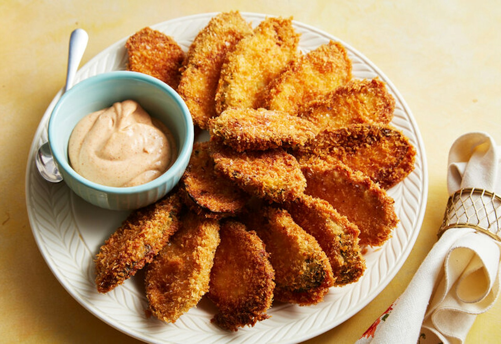 Fried zucchini chips with dish of smoked paprika aioli on white plate atop yellow surface.