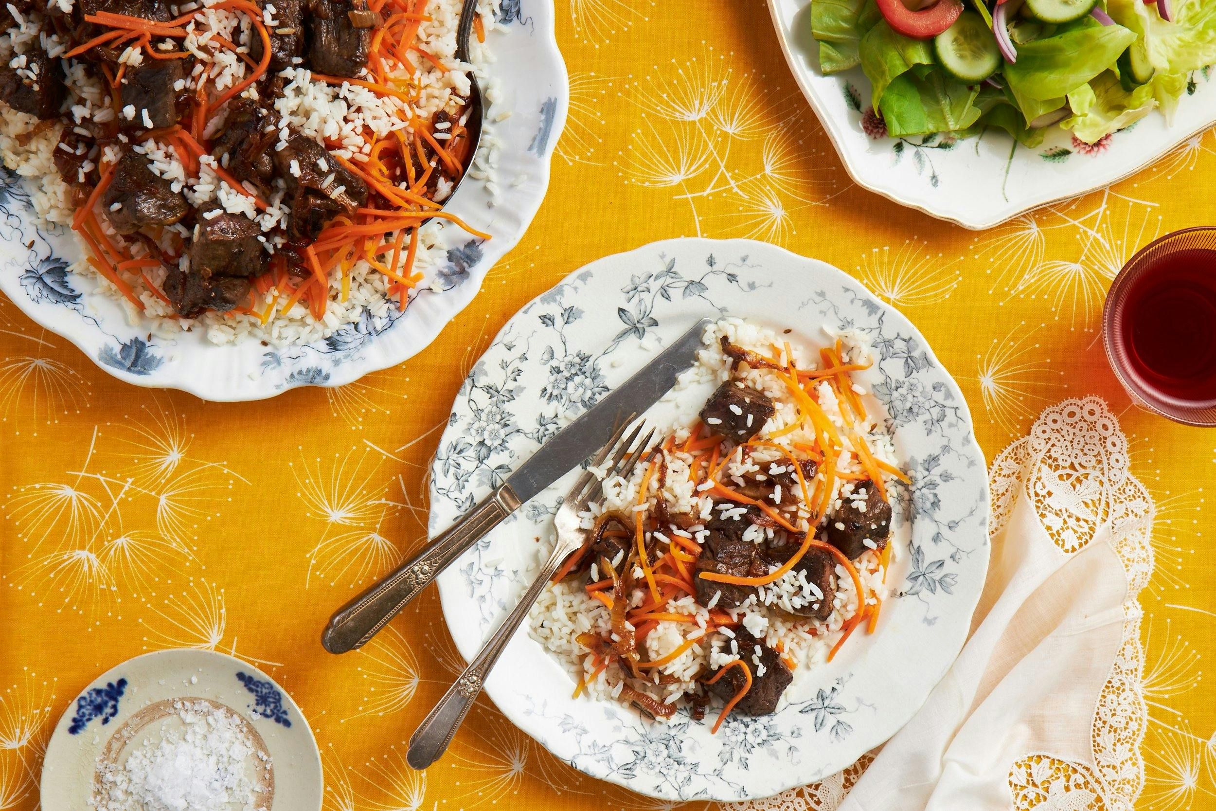 Plov with rice, beef, and carrots on white plate atop yellow tablecloth. 