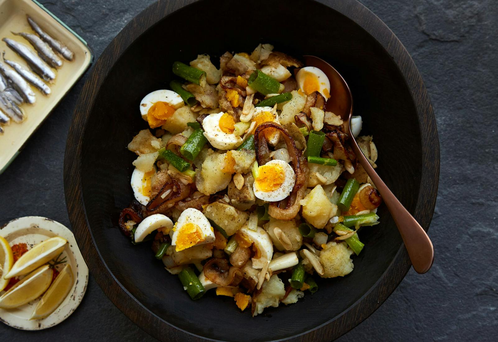 Eggs with onion in wooden bowl alongside plates of lemon wedges and anchovies, atop stone surface.