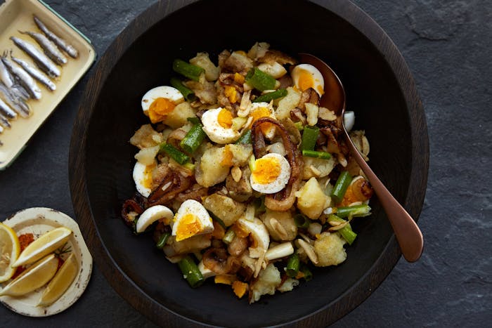 Eggs with onion in wooden bowl alongside plates of lemon wedges and anchovies, atop stone surface.