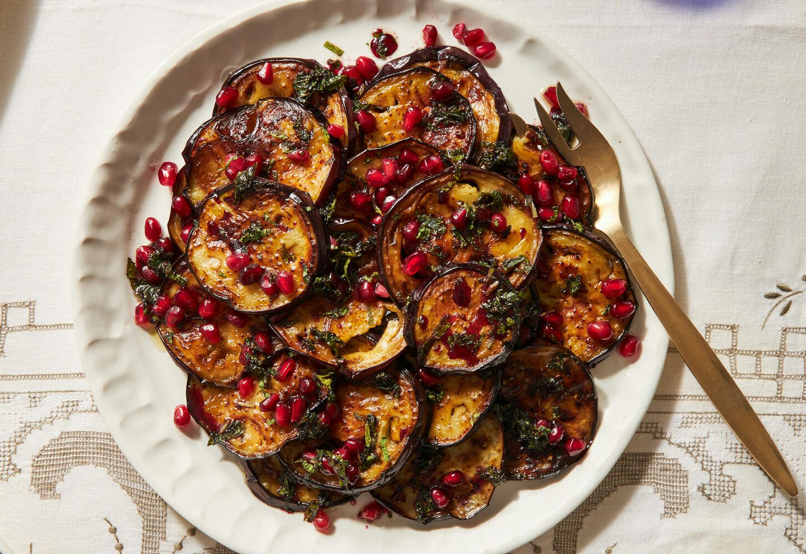 Fried eggplant with mint vinaigrette and pomegranate kernels on white plate atop white patterned tablecloth.