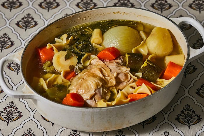 Chicken soup in white dutch oven atop white patterned tablecloth.
