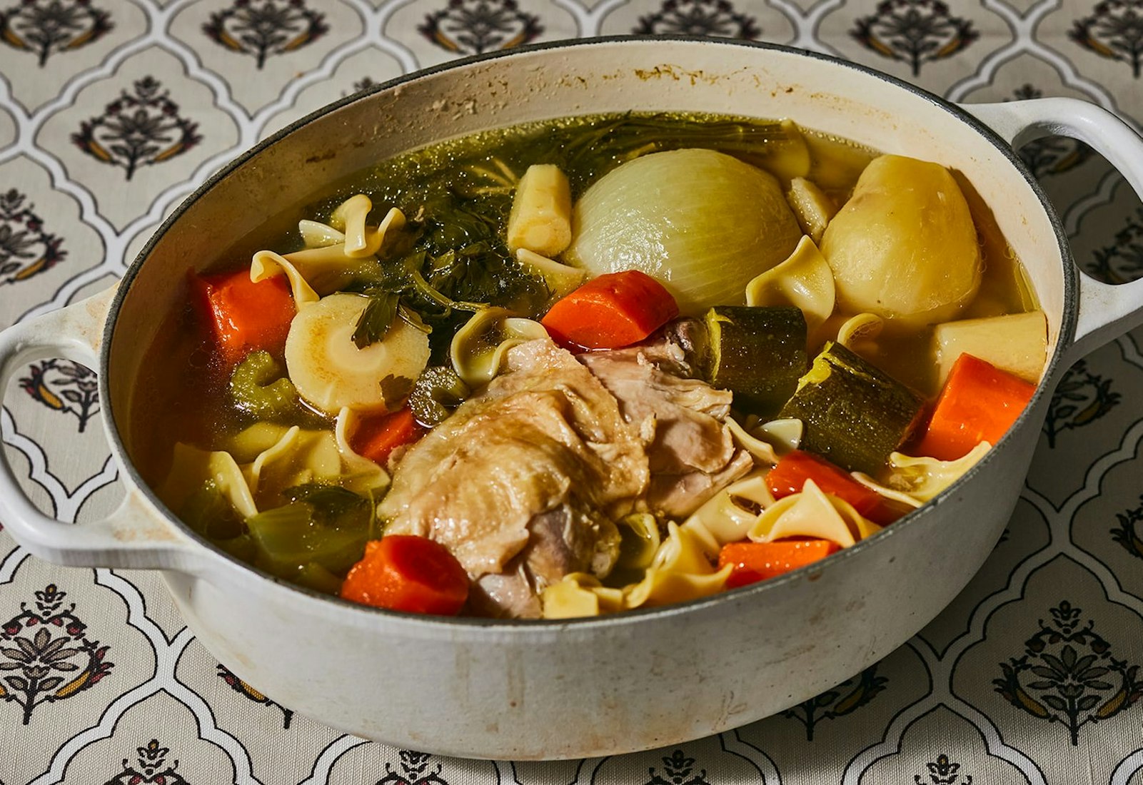 Chicken soup in white dutch oven atop white patterned tablecloth.