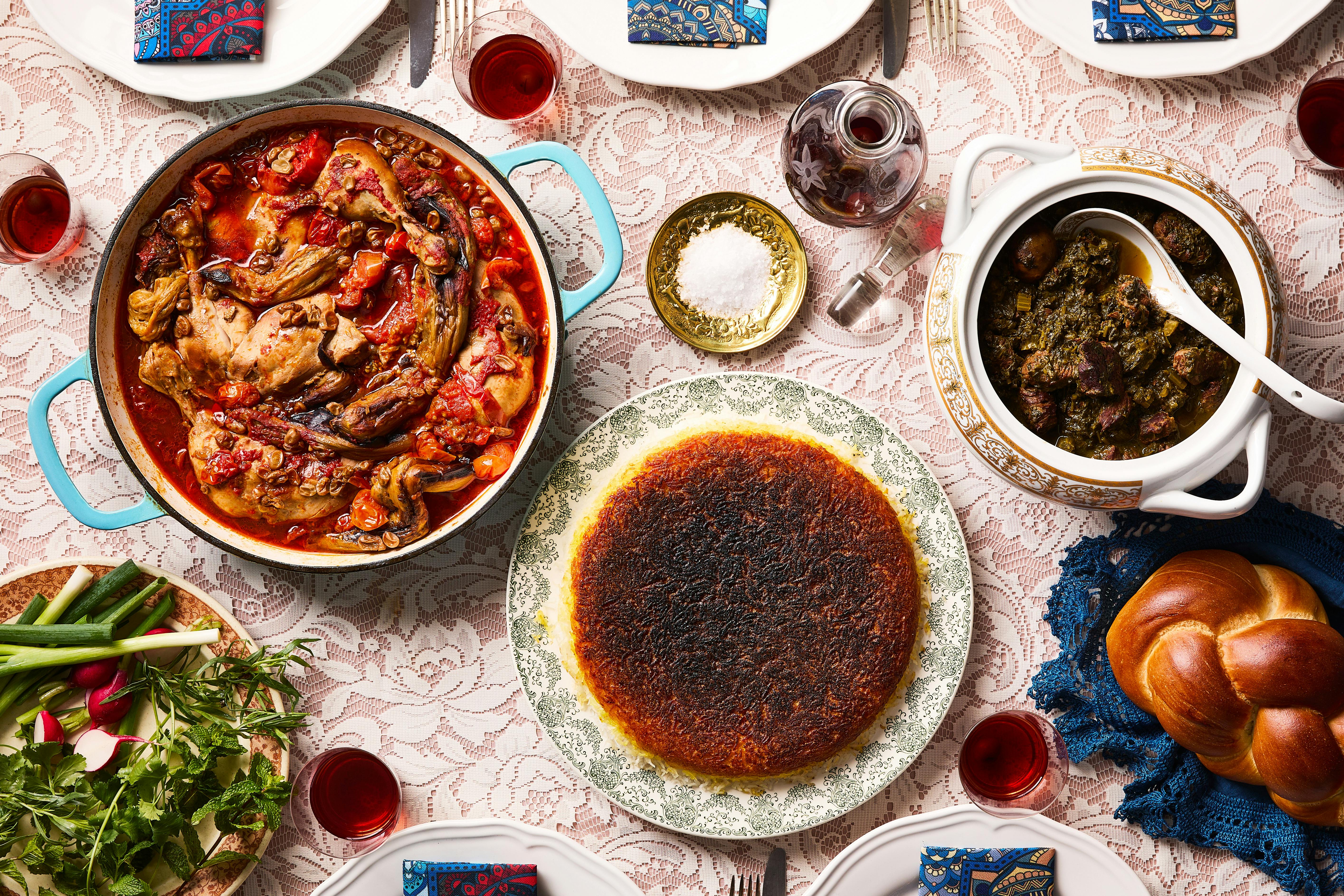 A light pink tablecloth is covered with dishes of joojeh bademjan, karafs khoresht, tahdig and shots of sour cherry vodka. 