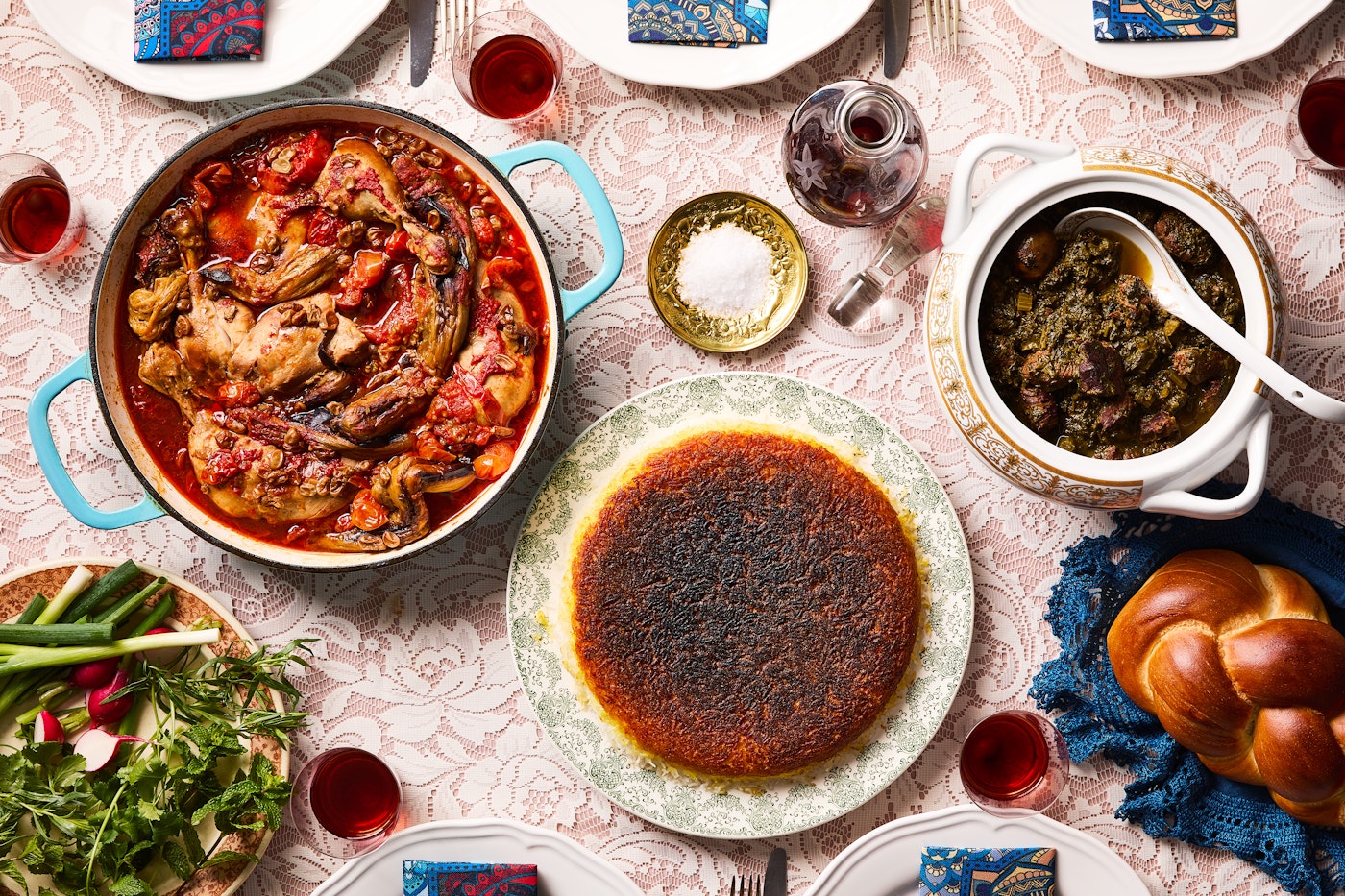 A light pink tablecloth is covered with dishes of joojeh bademjan, karafs khoresht, tahdig and shots of sour cherry vodka.