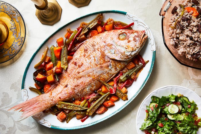 Whole steamed red snapper served over pumpkin, okra, and carrots on serving platter next to rice and peas and a green salad.