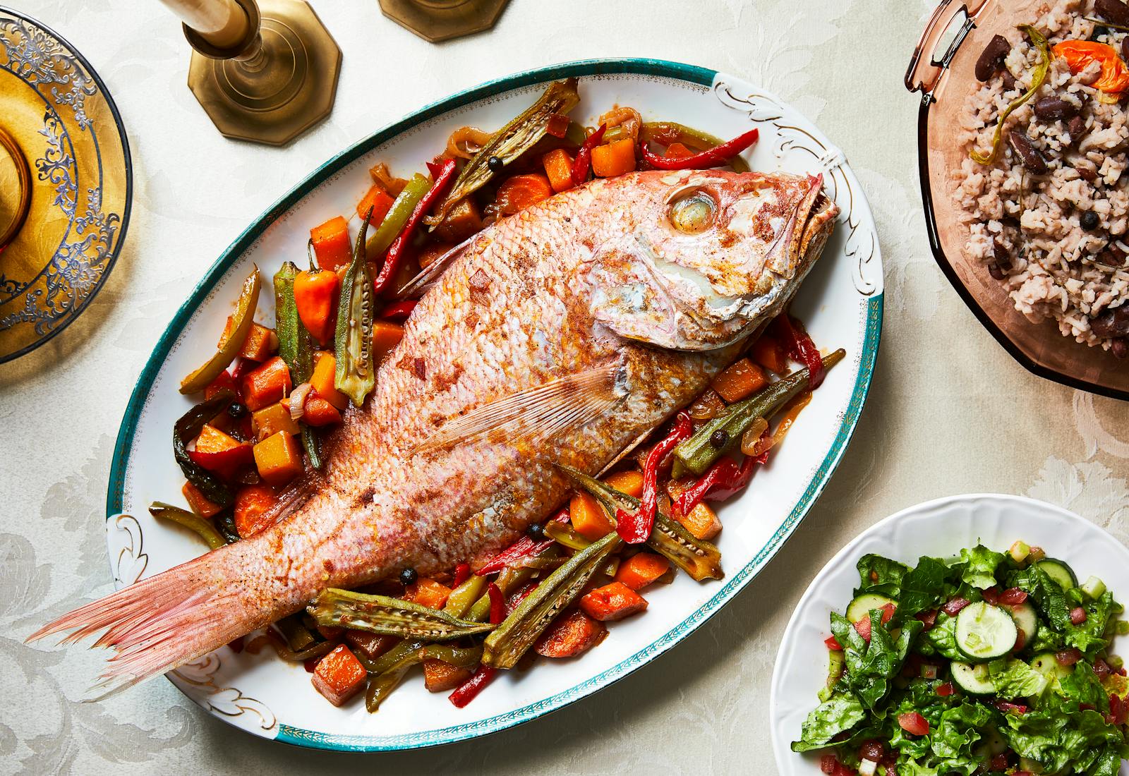 Whole steamed red snapper served over pumpkin, okra, and carrots on serving platter next to rice and peas and a green salad.