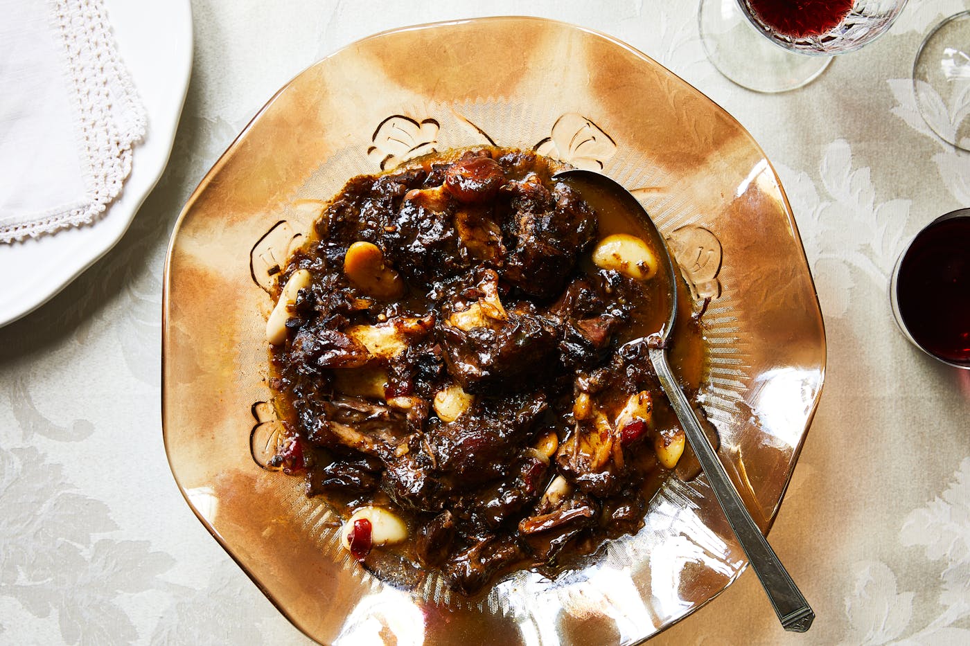 Oxtail stew served in large, glass amber bowl atop white floral tablecloth.
