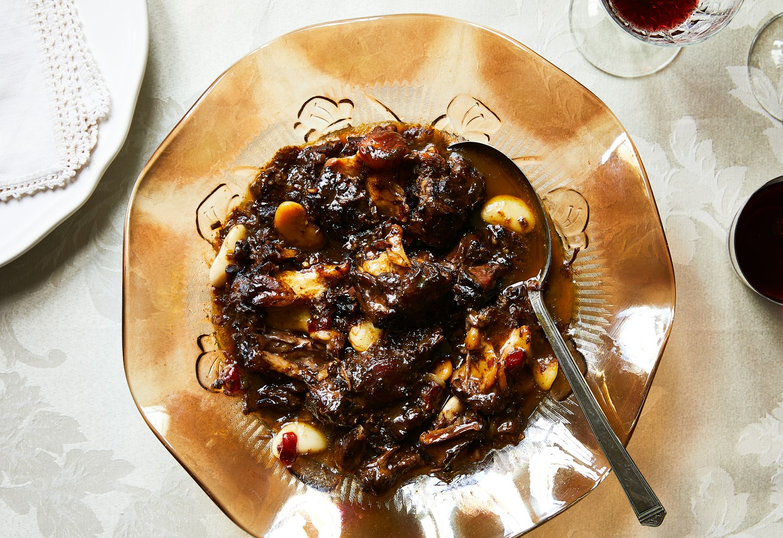 Oxtail stew served in large, glass amber bowl atop white floral tablecloth.