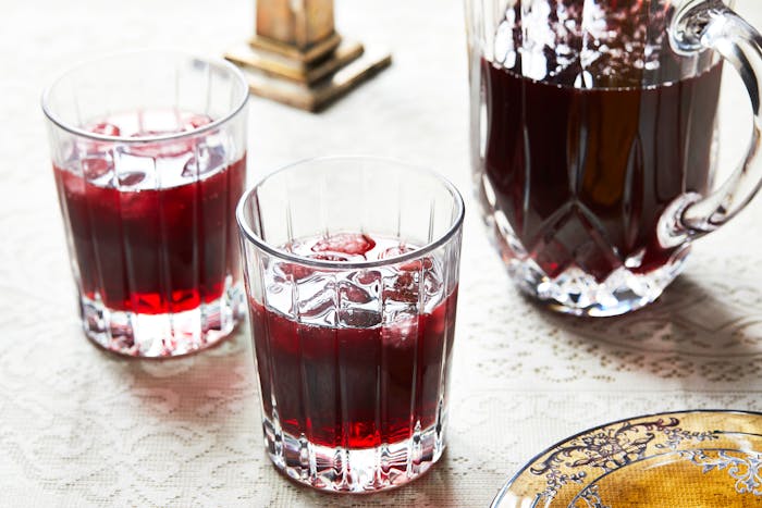 Two glasses of sorrel with ice next to pitcher of sorrel on lace tablecloth.