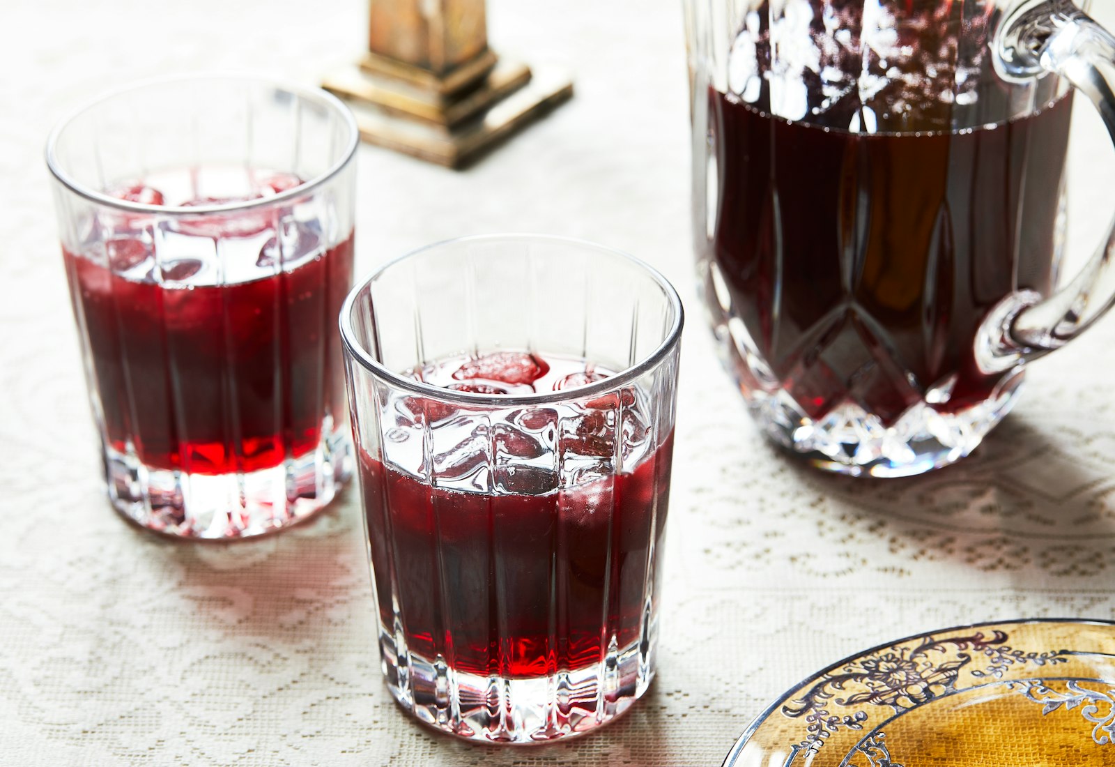 Two glasses of sorrel with ice next to pitcher of sorrel on lace tablecloth.