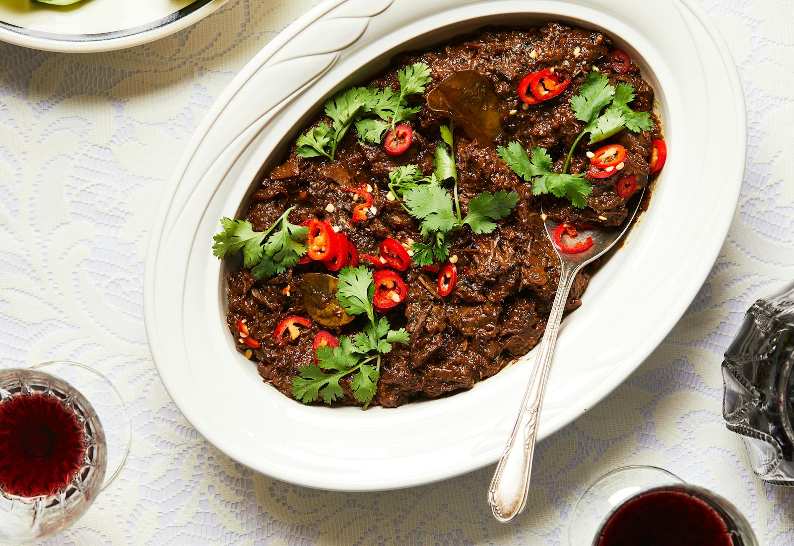 Beef rendang in white, oval serving bowl with wine in crystal glasses next to it.