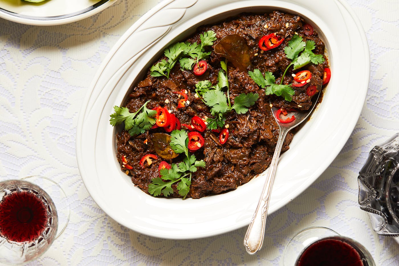 Beef rendang in white, oval serving bowl with wine in crystal glasses next to it.