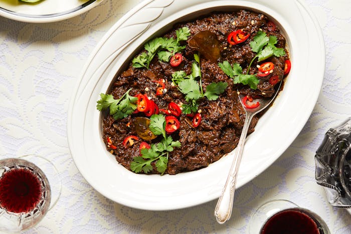Beef rendang in white, oval serving bowl with wine in crystal glasses next to it.