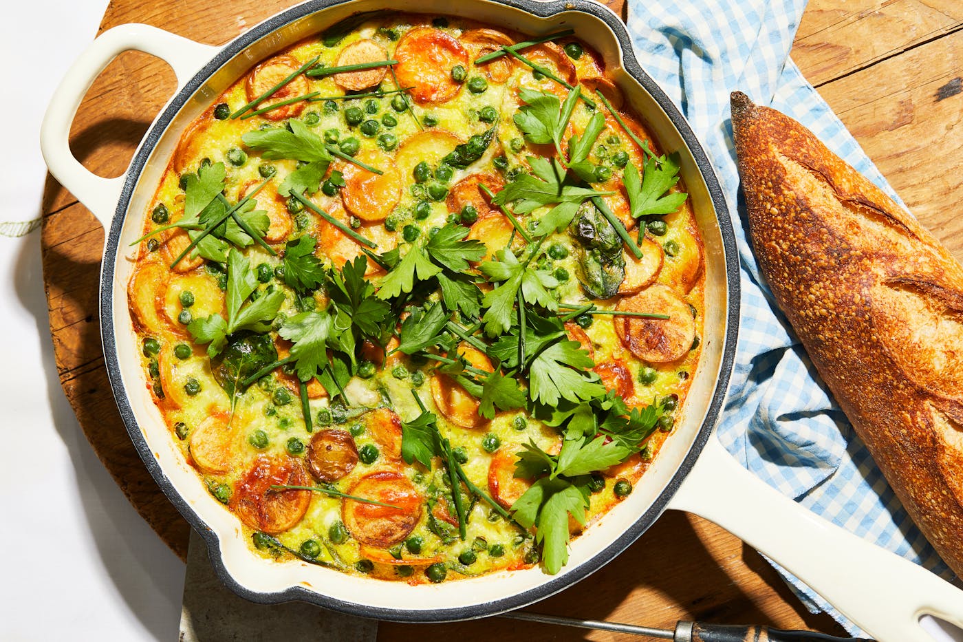 Algerian omelette in large, white cast iron pan over wooden cutting board beside a baguette.