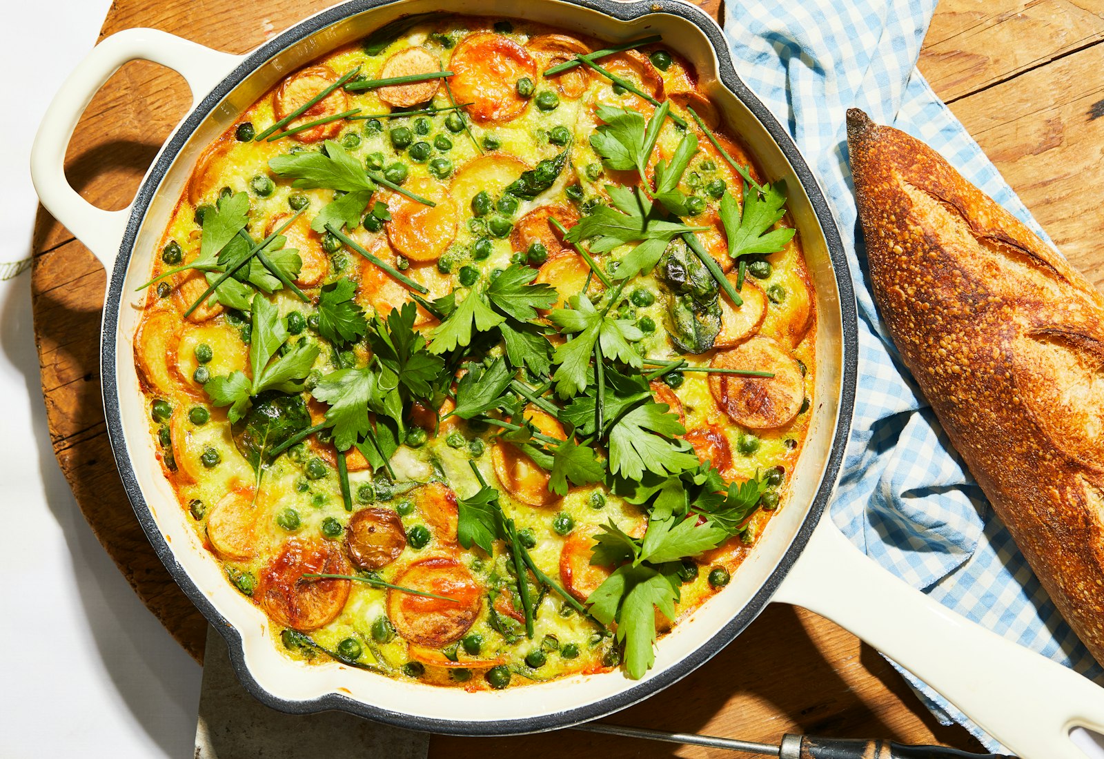 Algerian omelette in large, white cast iron pan over wooden cutting board beside a baguette.