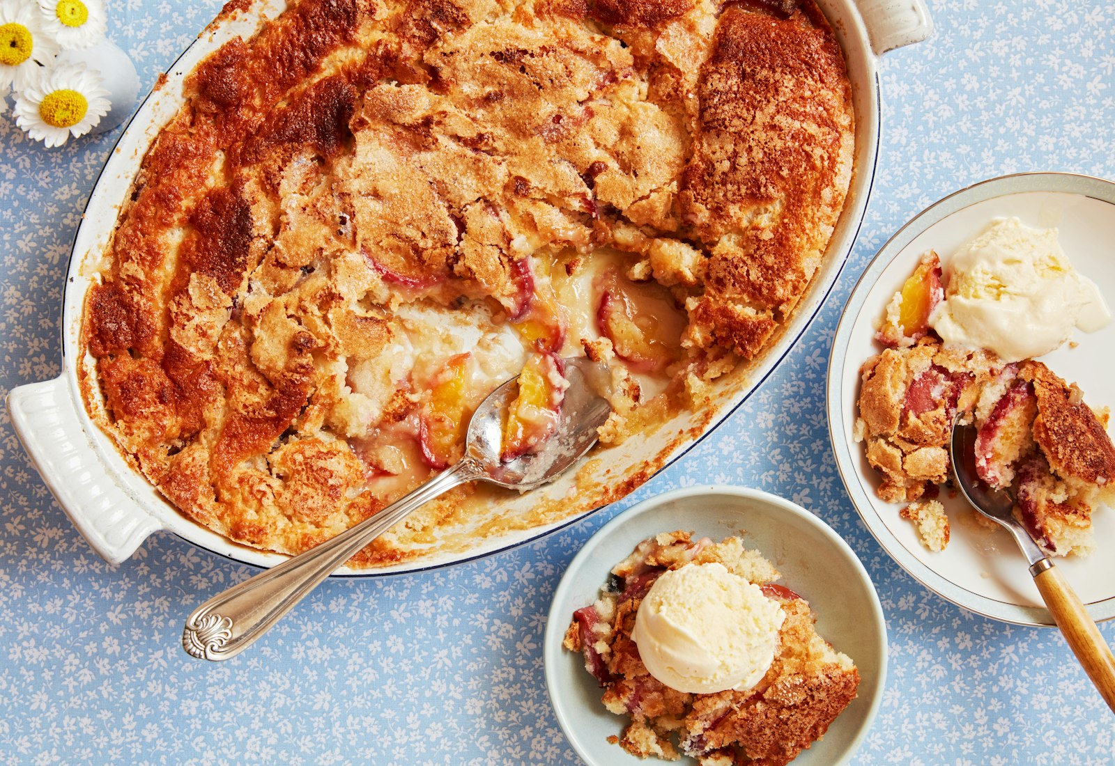 Peach cobbler in large, oval serving dish with a serving in one bowl and one plate beside it.