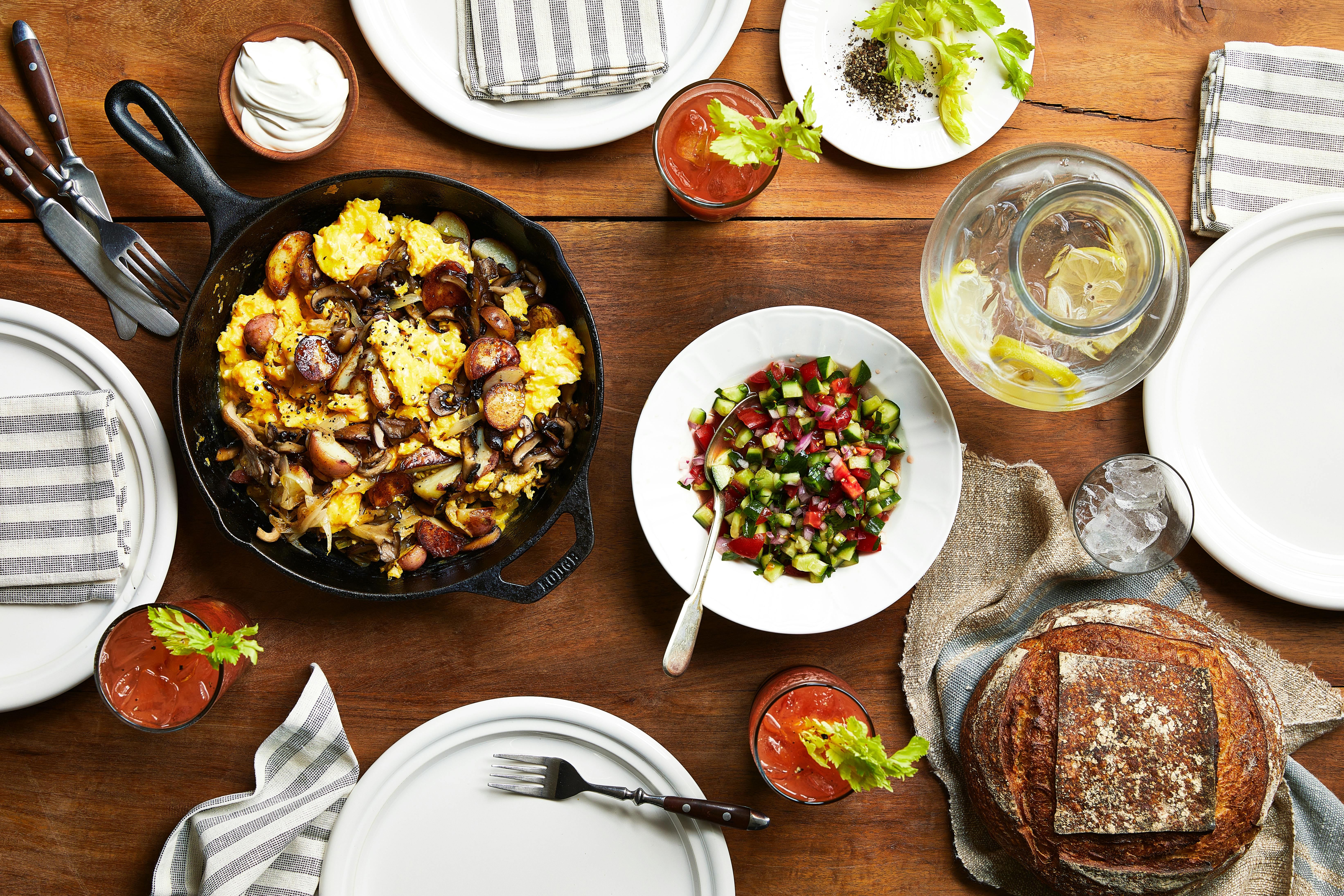 Tablespread with scrambled eggs with sauteed mushrooms and onions in cast iron, cucumber tomato salad, bloody Miriams and a loaf of bread.