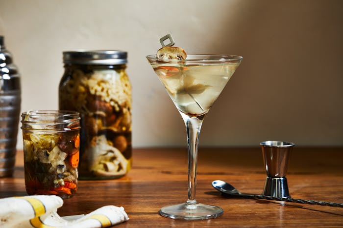 Pickled mushroom martini in martini glass atop dark wood table.