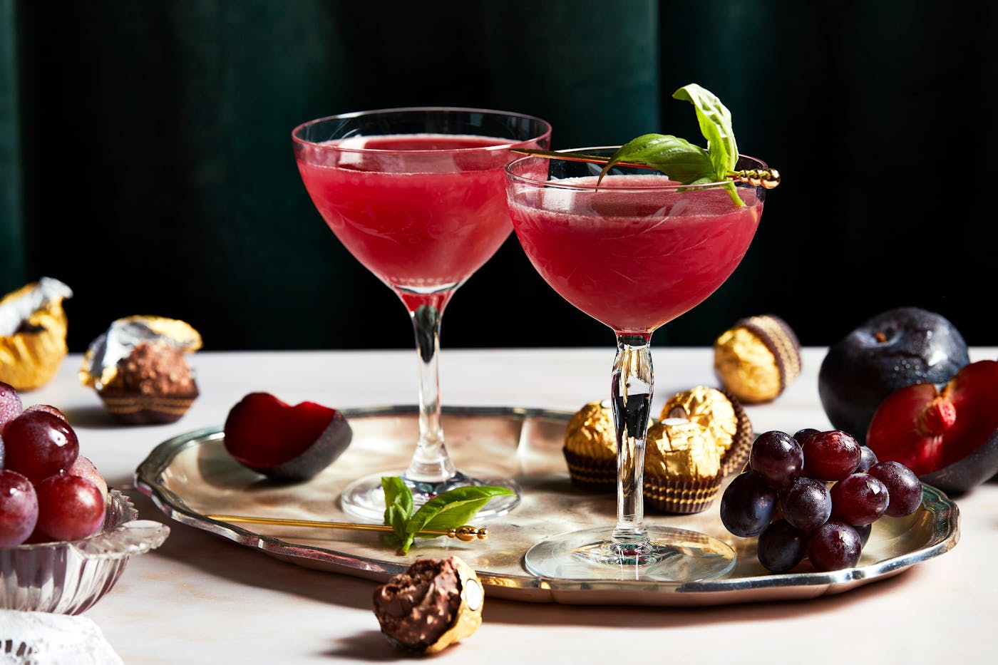 Ruby hued plum cocktails garnished with basil leaves stand against a dark green velvet background, on a tray with plums and Ferrero Rocher chocolates.