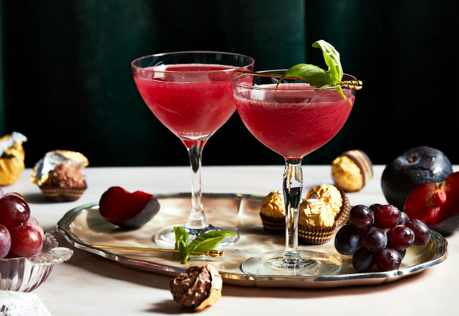 Ruby hued plum cocktails garnished with basil leaves stand against a dark green velvet background, on a tray with plums and Ferrero Rocher chocolates.