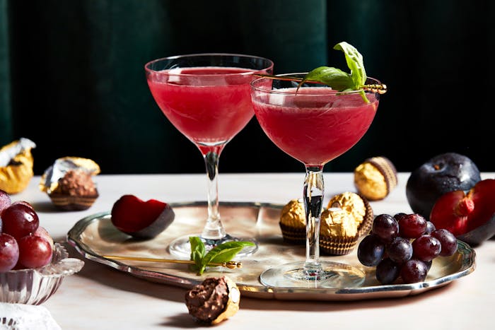 Ruby hued plum cocktails garnished with basil leaves stand against a dark green velvet background, on a tray with plums and Ferrero Rocher chocolates.