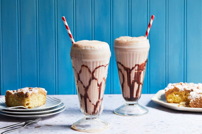 Chocolate Fizz Drinks in tall, egg-cream style glasses with red striped straws and crumb cake, against a blue background.