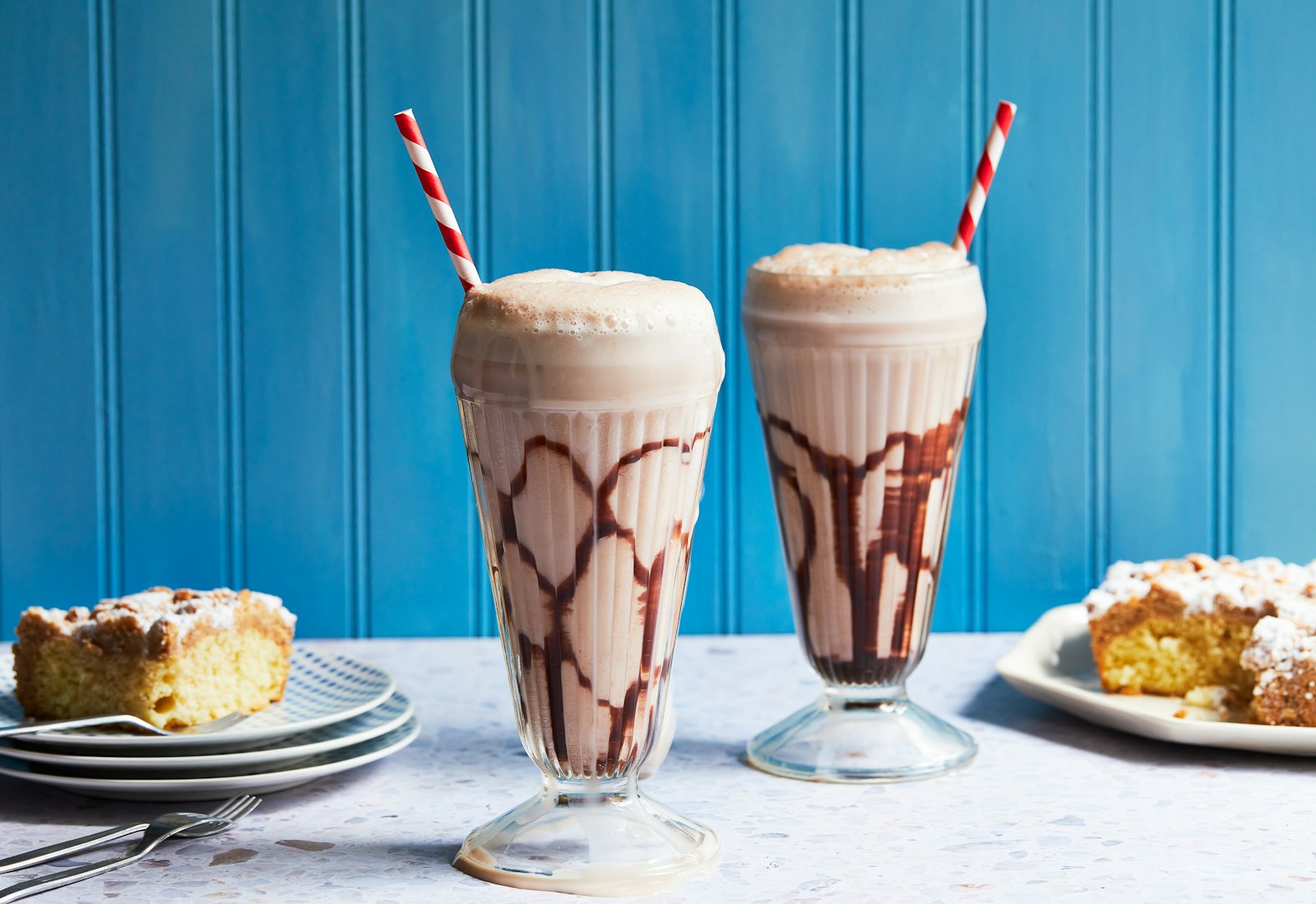 Chocolate Fizz Drinks in tall, egg-cream style glasses with red striped straws and crumb cake, against a blue background.