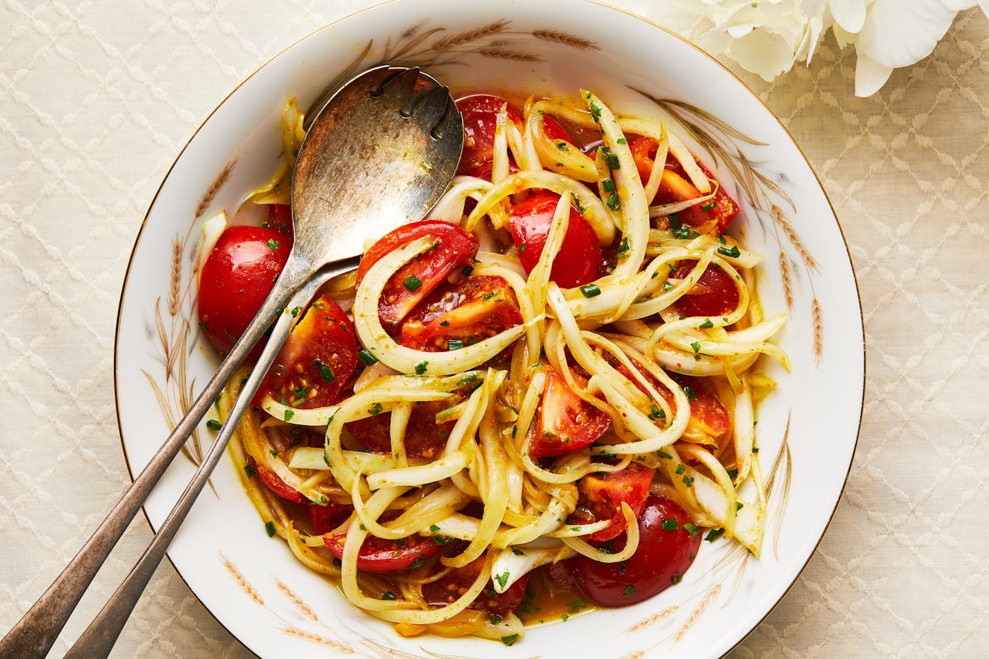 A salad of tomatoes and onions with amba dressing in a white bowl, with silver tongs.
