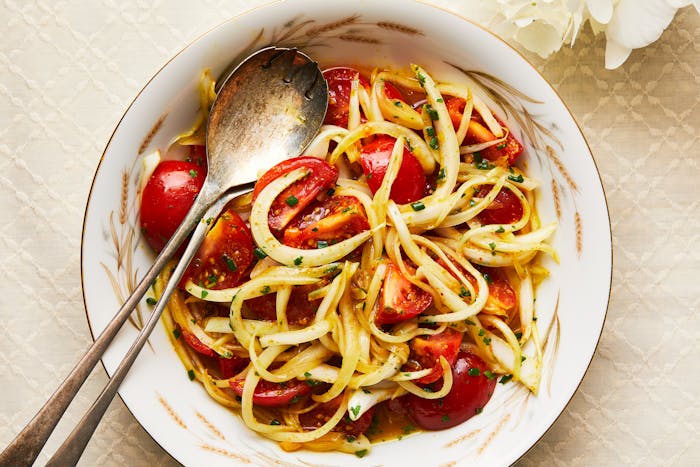 A salad of tomatoes and onions with amba dressing in a white bowl, with silver tongs.