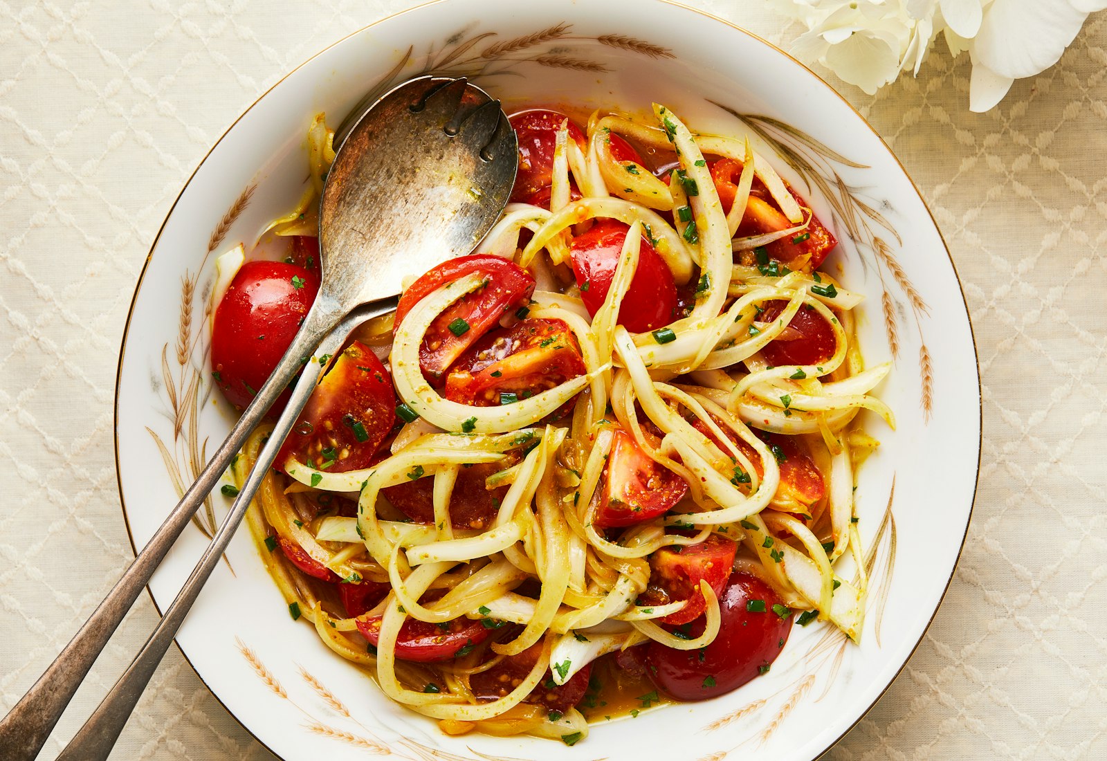 A salad of tomatoes and onions with amba dressing in a white bowl, with silver tongs.