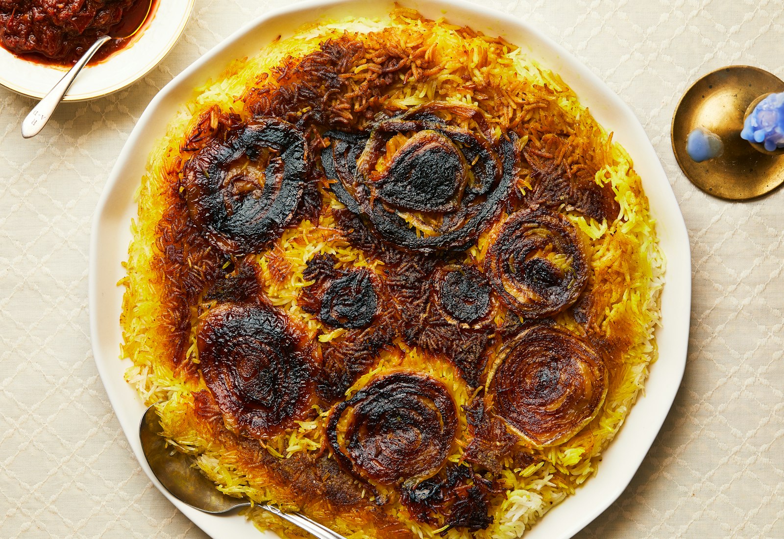 Spiced yellow rice featuring a layer of crispy onions on top, with a small bowl of matbucha and a candle in the corner.