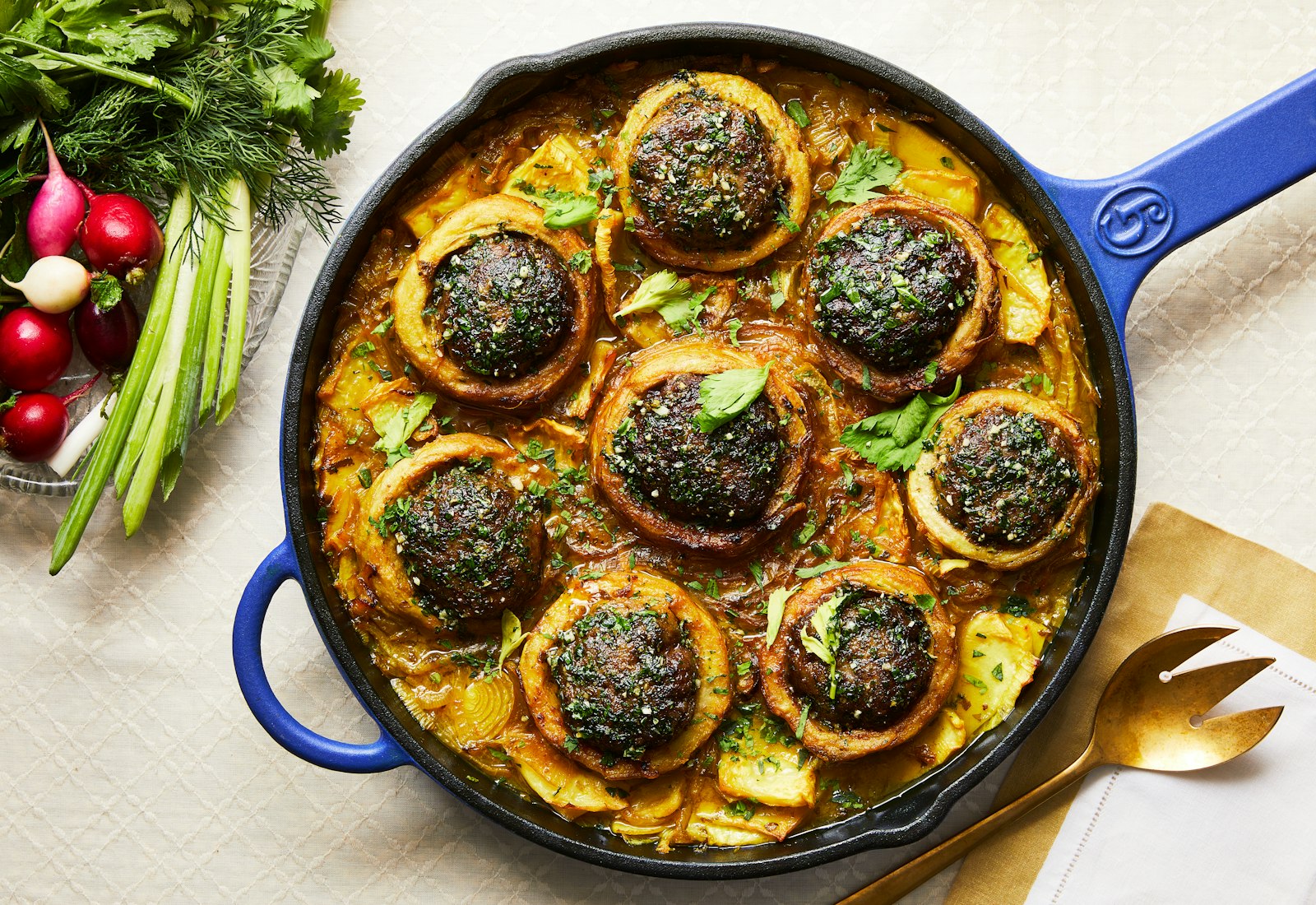 A bright blue pot filled with beef stuffed artichokes and lemon and celery sits on the table with a plate of vegetables in the corner and a gold fork. 