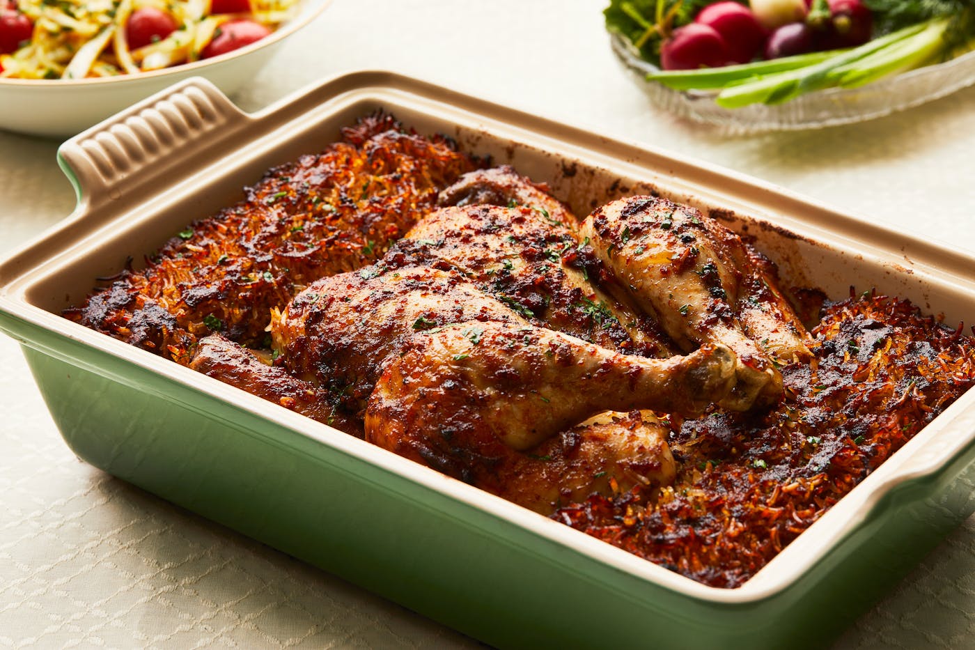 A green casserole dish filled with a whole, spiced chicken and crispy rice, with tomato salad and fresh radishes visible in the background. 