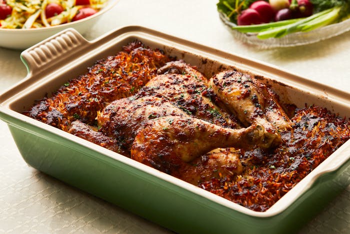 A green casserole dish filled with a whole, spiced chicken and crispy rice, with tomato salad and fresh radishes visible in the background. 