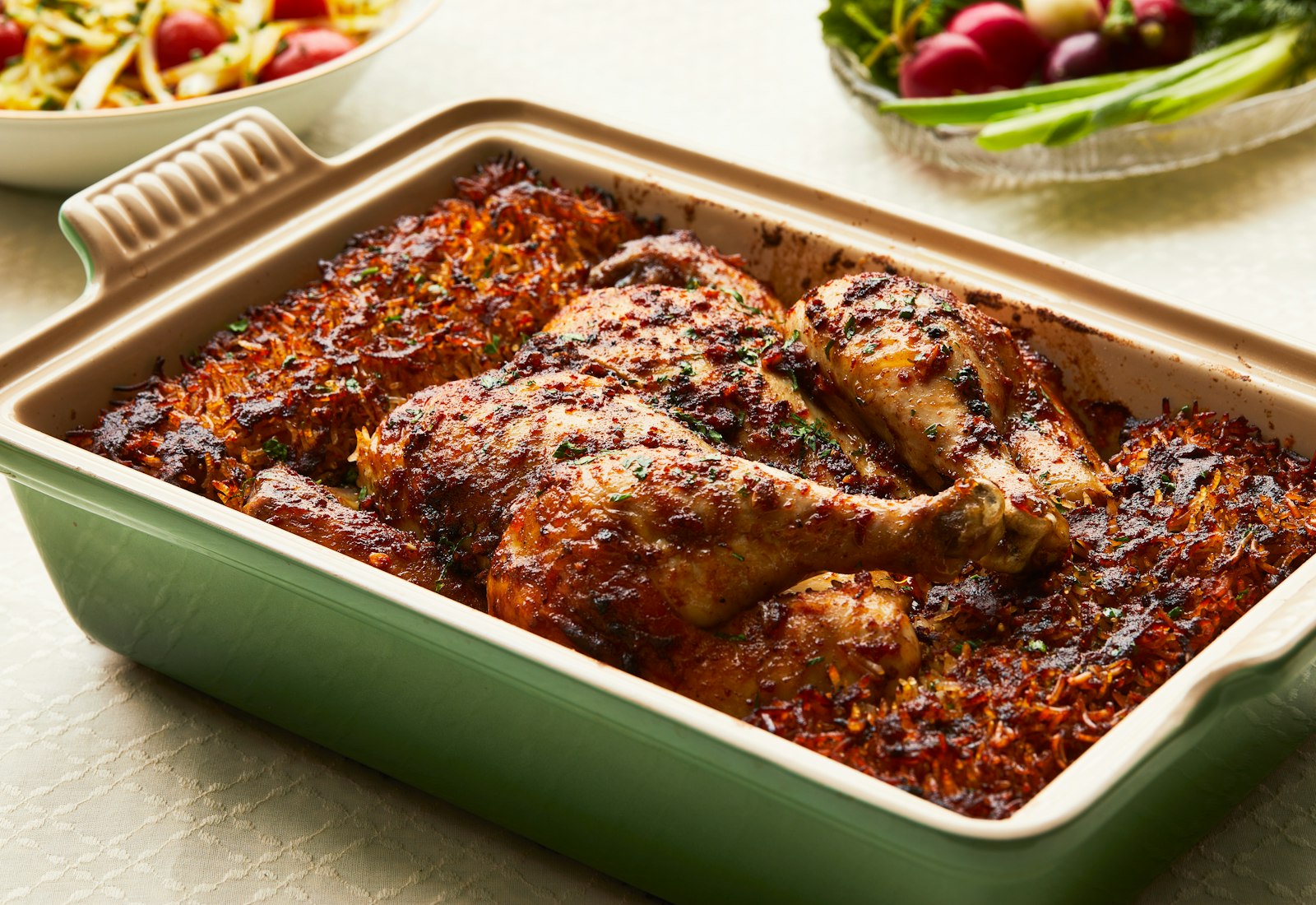 A green casserole dish filled with a whole, spiced chicken and crispy rice, with tomato salad and fresh radishes visible in the background.