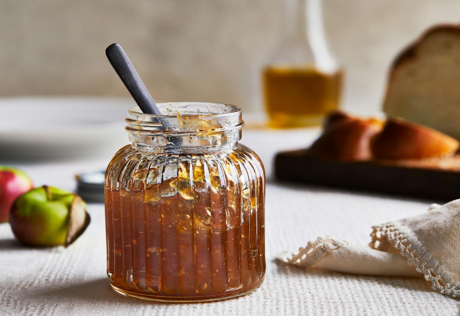 A glass jar of golden apple jam sits on a table with apples and challah in the background.