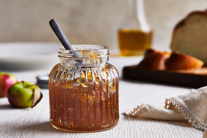A glass jar of golden apple jam sits on a table with apples and challah in the background.